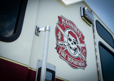 White emergency vehicle side with a red Hannibal Fire Department emblem featuring a skull and crossed axes beside a chrome latch and window edge