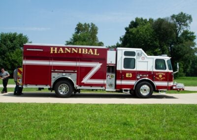 Red fire engine with Hannibal lettering parked on a sunny day, two people standing nearby on a grassy area with trees in the background.