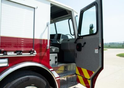 Open passenger door of red fire truck, showing the cabin interior and steps with yellow-and-red reflective stripes nearby.