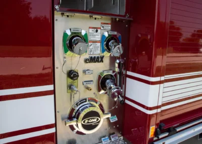 Fire engine control panel with chrome pumps, levers, and warning labels mounted on metal plate beside red exterior.