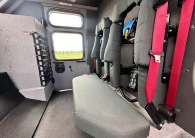Interior of a compact vehicle or cabin with gray seats and bright red seatbelts, window view of fields outside, and equipment racks along the wall.