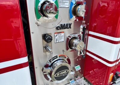 Close-up of a fire truck pump panel showing chrome valve knobs, gauges, and labeled discharges and intake on a red vehicle side.