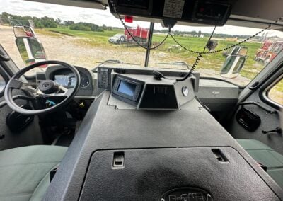Inside view of a truck cab showing the steering wheel, dashboard screen, and beaded microphone cord, with a grassy lot and red trailers outside.