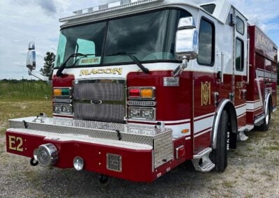 Red fire truck parked on a gravel lot, front view with chrome grille and emergency lights on top, Macon Fire Dept. on the cab.