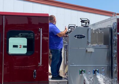 Firefighter in a blue shirt standing on a metal platform beside a silver fire pump panel on a red fire truck, water spraying from hoses nearby