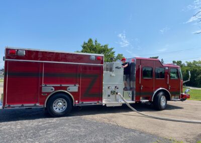 Red fire truck with a long hose on the pavement and a firefighter in a red cap standing by on a sunny day.