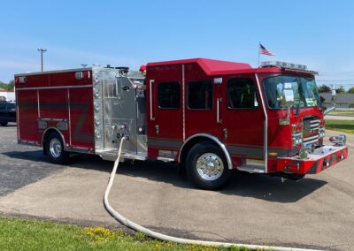 Red fire engine with a long beige hose laid across a paved lot under a clear blue sky, American flag visible on top.