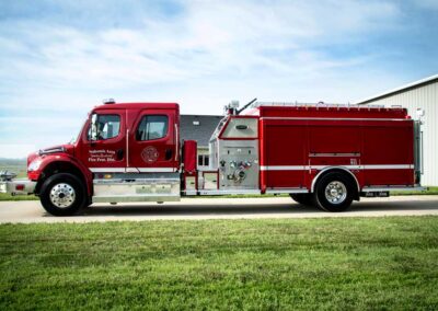 Red fire engine parked on a concrete path beside a grassy field and a light industrial building in the background.
