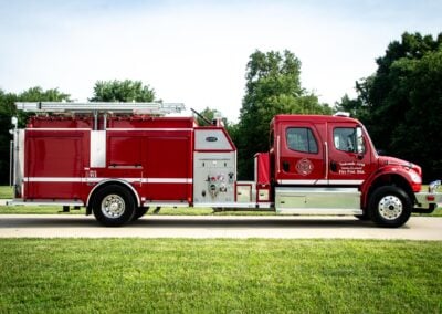 Red fire truck parked on a grassy field with trees in the background on a sunny day, side view showing equipment compartments and ladder rack.