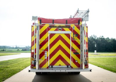 Rear view of a fire engine with yellow-and-red chevron markings, equipment bays visible, and a ladder mounted on top, parked on a sunny street.