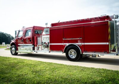 Red fire truck parked on a paved road with grass and trees nearby under a bright sky.