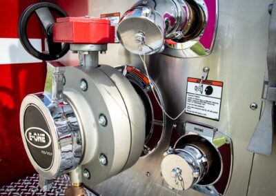 Close-up of industrial pump components: red valve, chrome fittings, and warning labels on a metal panel