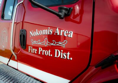 Close-up of a red fire truck door with 'Nokomis Area Fire Prot. Dist.' lettering and emblem visible on the side.