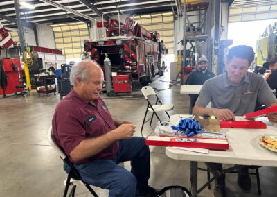 Fire station bay with red fire trucks in the background; people sit at folding tables, wrapping gifts and eating pizza.
