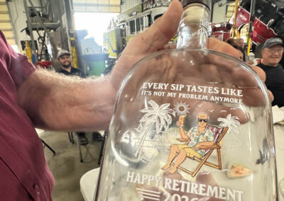Close-up of a glass keepsake with a tropical scene and the words 'EVERY SIP TASTES LIKE' and 'HAPPY RETIREMENT 2026'; hand holding it in a workshop with people in the background.