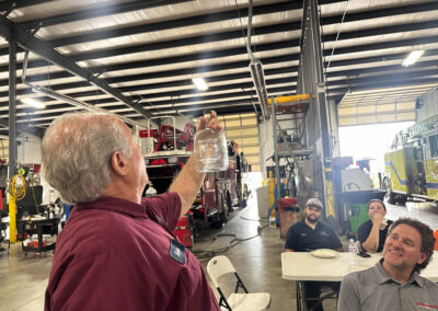 Man in a maroon shirt holding up a clear bottle as if to toast, in a busy workshop with coworkers at tables and equipment visible in the background.