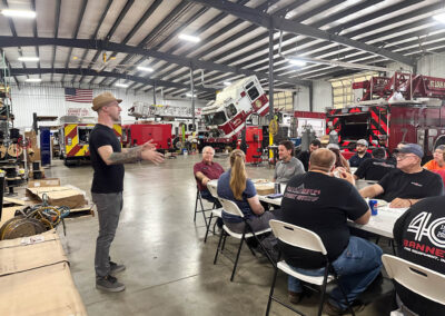 A man in a hat speaks to a seated group inside a fire station workshop, with firetrucks and equipment in the background.