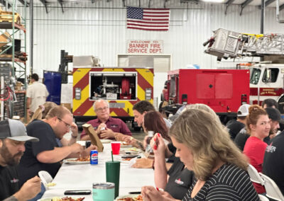 People seated at a long cafeteria-style table inside a fire station, eating meals with red cups and drinks, firefighters in background, American flag overhead.