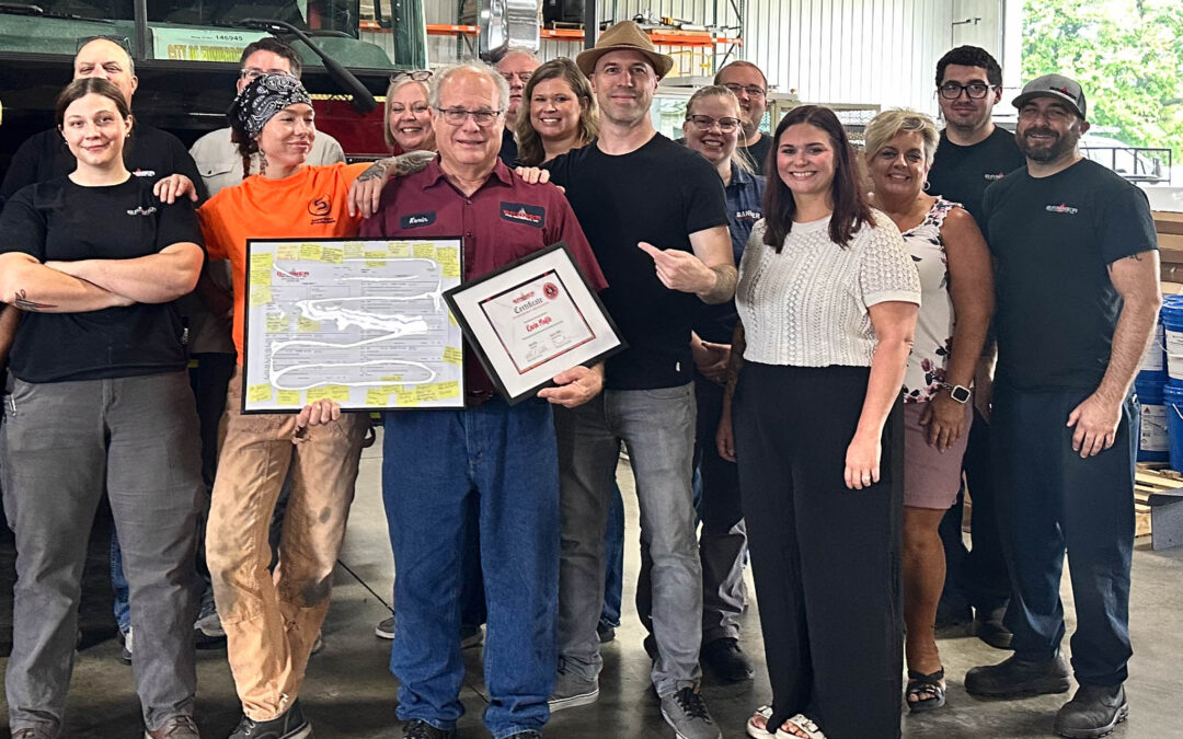 Group of coworkers in a workshop posing for a photo, two men in the front hold framed certificates while others smile beside them.