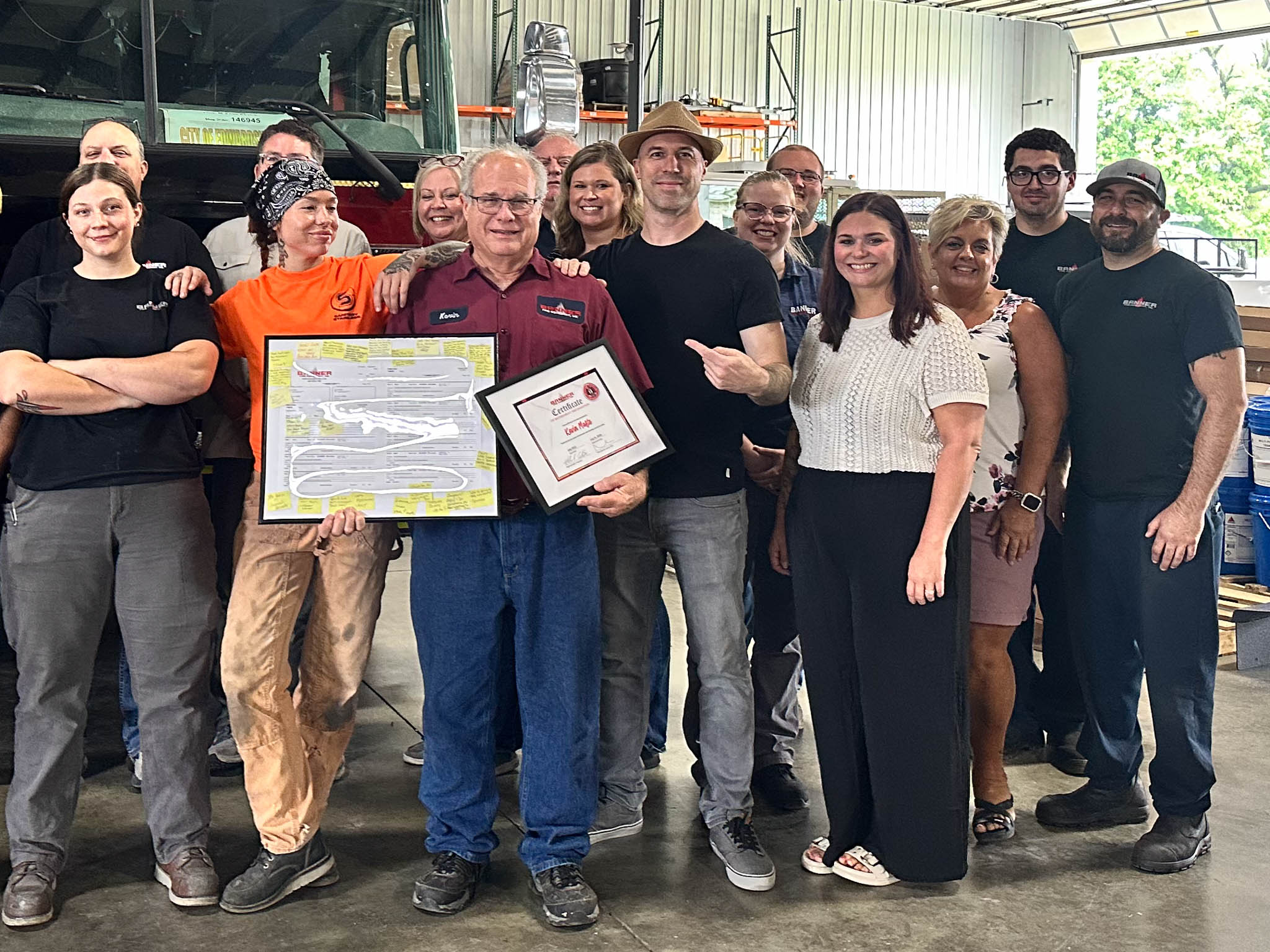 Group of coworkers in a workshop posing for a photo, two men in the front hold framed certificates while others smile beside them.