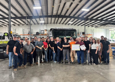 Group of smiling workers posing in a garage workshop with a large truck behind them, indoors.