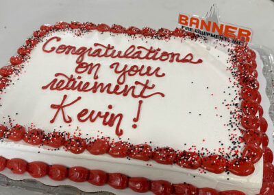 Cake celebrating retirement with red script reading 'Congratulations on your retirement' and a red rosette border with sprinkles, plus a 'BANNER FIRE EQUIPMENT' topper (informational).