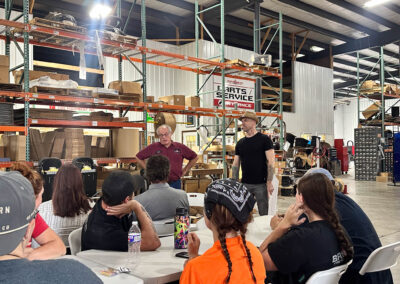 Group of people seated at tables in a warehouse, listening to two men speaking at the front.