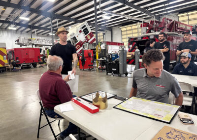 Fire station workshop: people seated at a table with paperwork while red fire engines and equipment line the background.
