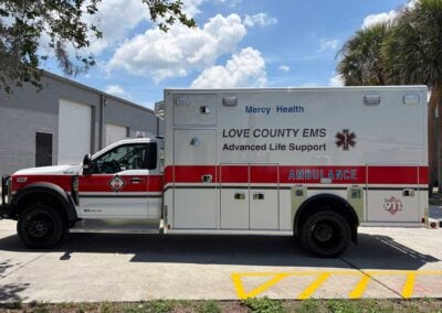 Side view of a white and red Mercy Health ambulance with Love County EMS branding in a parking lot by a gray building beneath a blue sky with palm trees.