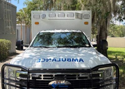 Front view of a white ambulance with a black grille guard, roof emergency lights, and 'AMBULANCE' text reflected on the hood under trees and foliage.