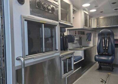 Interior of a modern ambulance with stainless steel cabinets, medical equipment, and blue captain chairs for crew.