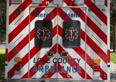 Rear view of a red-and-white chevron ambulance with Mercy Health branding, two rear doors, and a Florida license plate (EBF3421). They read 'LOVE COUNTY AMBULANCE' on the back.