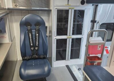 Ambulance interior with a blue seated patient chair, seat belts, and storage cabinets on the wall behind it.