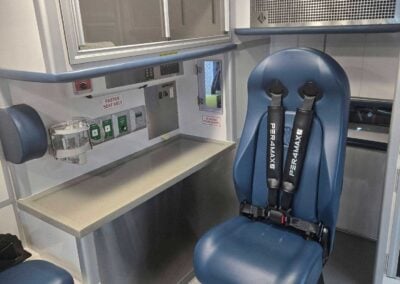 Interior of a medical transport vehicle with a blue single-seat stretcher chair and overhead medical equipment panels along the wall, including a 'FASTEN SEAT BELT' sign and oxygen supplies nearby.