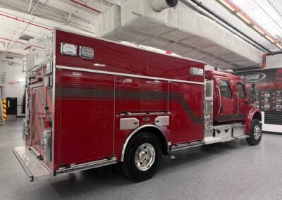 Red fire engine parked indoors in a maintenance bay with exposed HVAC ducts overhead, side compartments closed, and chrome wheels visible.
