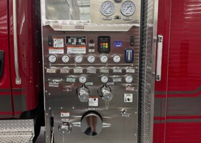 Fire truck control panel with multiple gauges, valves, and switches mounted on a metallic panel in a red emergency vehicle bay door area