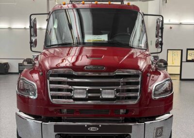 Front view of a red Freightliner semi-truck indoors, showing chrome grille, headlights, and large side mirrors in a showroom-like garage.