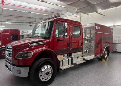 Red fire engine in a maintenance bay, showing chrome bumper, large tires, and equipment compartments on the side.