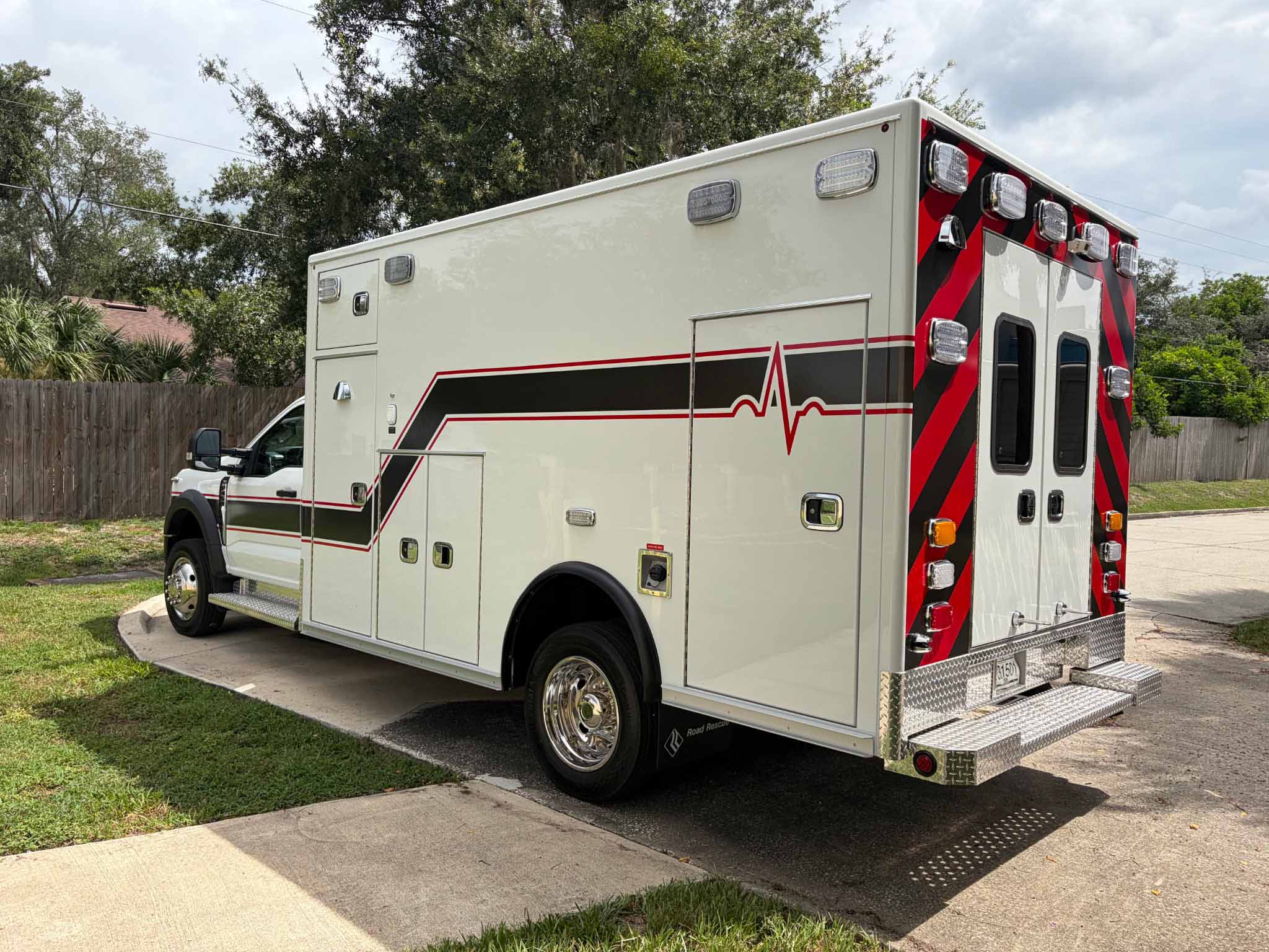 734112_EMS-2 Rear and side view of a white emergency ambulance with red and black chevron stripes and multiple roof lights, parked on a street.
