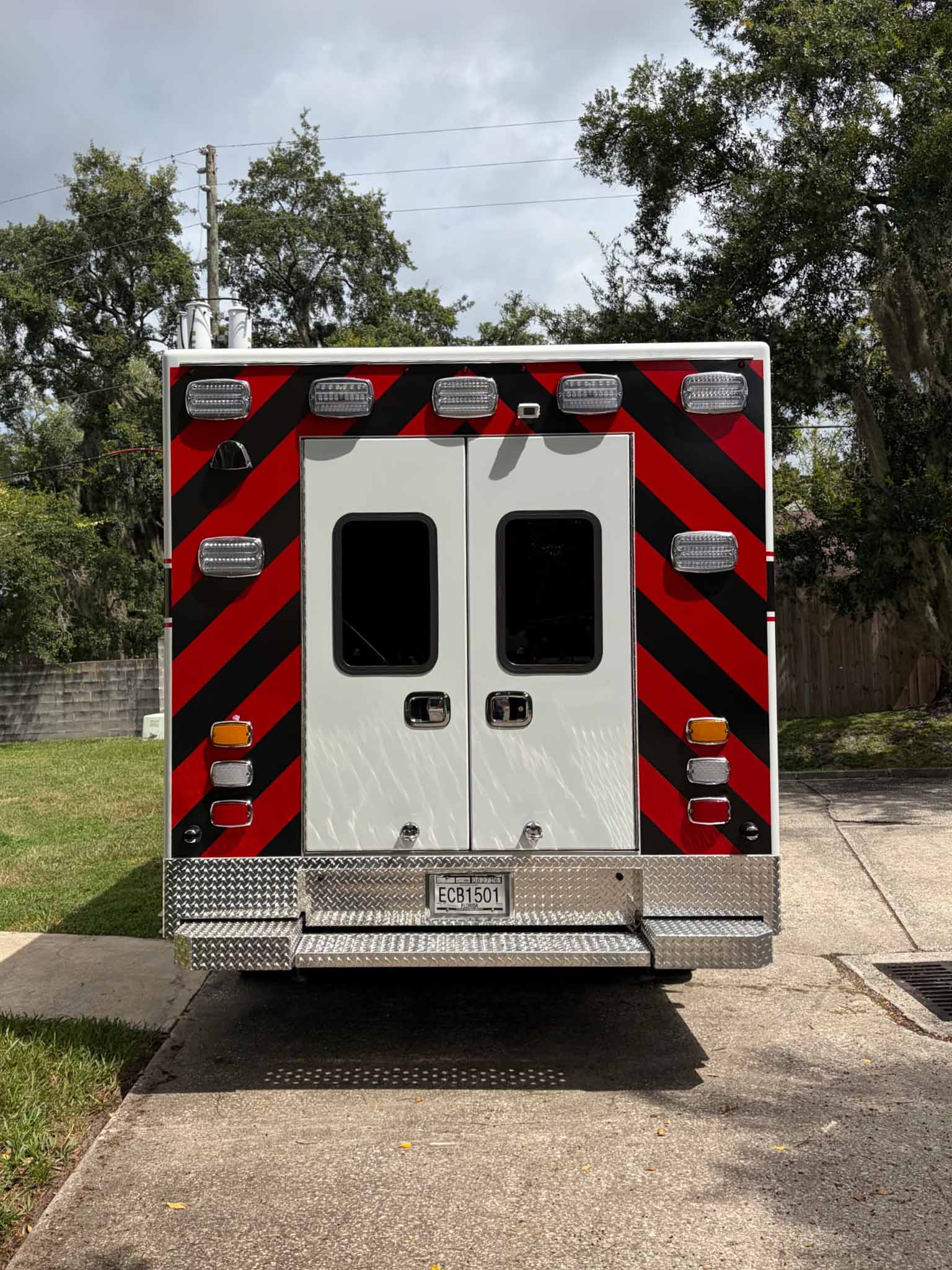 734112_EMS Rear and side view of a white emergency ambulance with red and black chevron stripes and multiple roof lights, parked on a street.