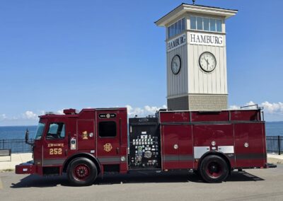 Red fire engine parked by the water next to a white clock tower with 'HAMBURG' signs against a clear blue sky.