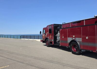 Red fire engine parked beside a waterfront with a blue sky and a black railing along the ocean behind it.