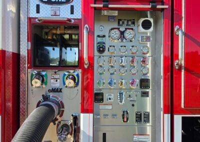 Close-up of a red fire truck pump panel with multiple gauges, levers, and a large hose connected at the bottom left.
