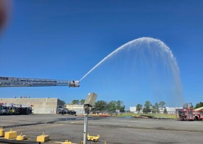 Industrial yard scene with a tall ladder truck spraying a high arc of water into the blue sky; construction equipment and buildings in the background.