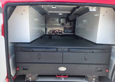 Interior of a red vehicle showing a battery box and wiring mounted to metal panels in the rear cargo area