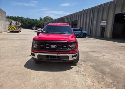 Red Ford truck in the foreground of an industrial yard, with a large warehouse on the right and a fire truck parked along the left side; clear blue sky overhead.