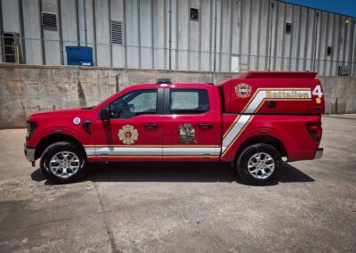 Red fire department pickup truck with Battalion 4 markings parked along a concrete barrier in front of a tall industrial building with vents and a chain-link fence behind it.