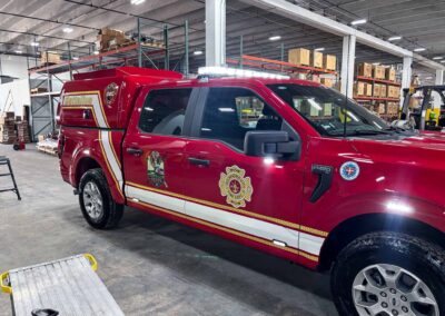 Red fire department pickup parked inside a warehouse with pallet racks and cardboard boxes on the shelves behind it, gear visible on top of the truck cab with emergency lights on top.