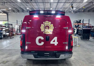 Rear view of a red fire department rescue vehicle with Oklahoma City Fire Dept. emblem, inside a workshop with shelves and equipment visible in the background.