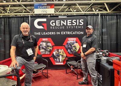 Two Genesis Rescue Systems representatives pose at a trade show booth with a large banner reading 'GENESIS RESCUE SYSTEMS' behind them.